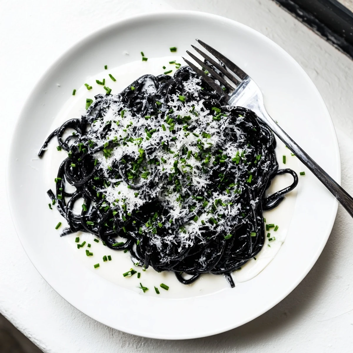 Glossy black squid ink pasta twirled in a velvety cream sauce, garnished with fresh chives and lemon zest on a white plate.  