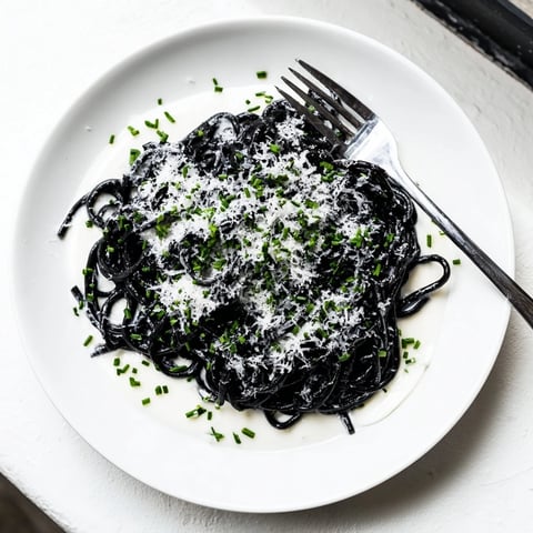 Glossy black squid ink pasta twirled in a velvety cream sauce, garnished with fresh chives and lemon zest on a white plate.  