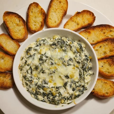 A close-up of Spinach Artichoke Dip, rich with cheese and ready for dipping toasted bread.