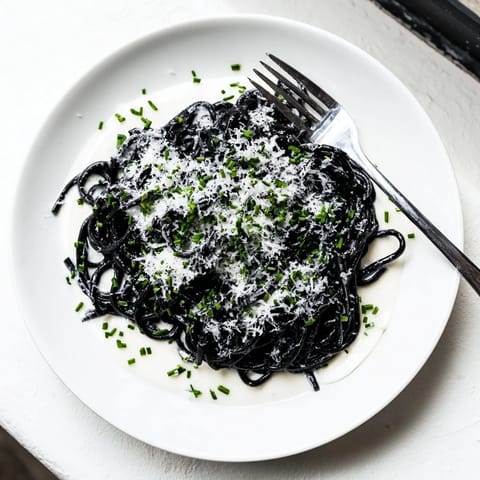 Glossy black squid ink pasta twirled in a velvety cream sauce, garnished with fresh chives and lemon zest on a white plate.  